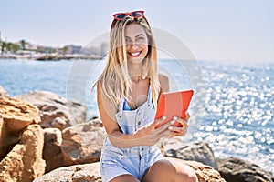Young blonde girl using touchpad sitting on the rock at the beach