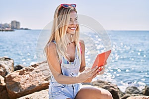 Young blonde girl using touchpad sitting on the rock at the beach