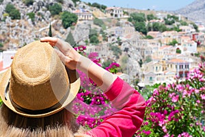 Young blonde girl with wavy hair in straw hat is watching on blooming oleander flowers and tiny colorful houses on greek island Sy
