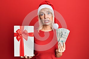 Young blonde girl wearing christmas hat, holding a gift and dollars clueless and confused expression