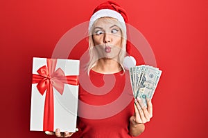 Young blonde girl wearing christmas hat, holding a gift and dollars making fish face with mouth and squinting eyes, crazy and