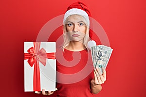 Young blonde girl wearing christmas hat, holding a gift and dollars relaxed with serious expression on face