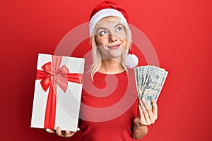 Young blonde girl wearing christmas hat, holding a gift and dollars smiling looking to the side and staring away thinking