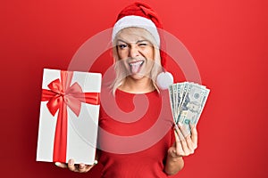 Young blonde girl wearing christmas hat, holding a gift and dollars sticking tongue out happy with funny expression