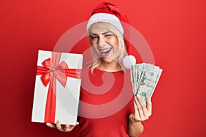 Young blonde girl wearing christmas hat, holding a gift and dollars winking looking at the camera with sexy expression, cheerful