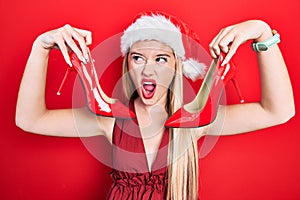 Young blonde girl wearing christmas hat holding high heel shoes angry and mad screaming frustrated and furious, shouting with