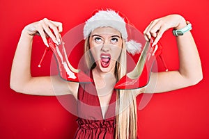 Young blonde girl wearing christmas hat holding high heel shoes angry and mad screaming frustrated and furious, shouting with