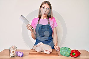 Young blonde girl wearing cook apron cooking chicken scared and amazed with open mouth for surprise, disbelief face