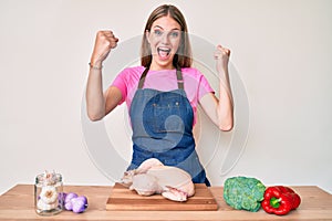 Young blonde girl wearing cook apron and holding chicken screaming proud, celebrating victory and success very excited with raised