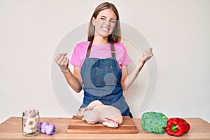 Young blonde girl wearing cook apron and holding chicken very happy and excited doing winner gesture with arms raised, smiling and