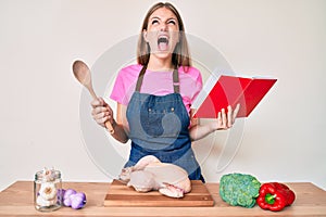 Young blonde girl wearing professional baker apron reading cooking recipe book angry and mad screaming frustrated and furious,