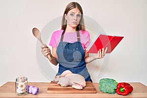 Young blonde girl wearing professional baker apron reading cooking recipe book clueless and confused expression