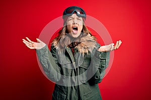 Young blonde girl wearing ski glasses and winter coat for ski weather over red background celebrating mad and crazy for success