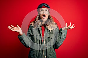 Young blonde girl wearing ski glasses and winter coat for ski weather over red background crazy and mad shouting and yelling with