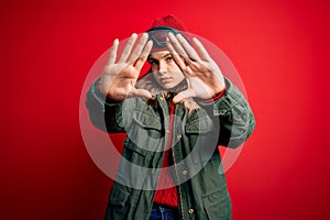 Young blonde girl wearing ski glasses and winter coat for ski weather over red background doing frame using hands palms and