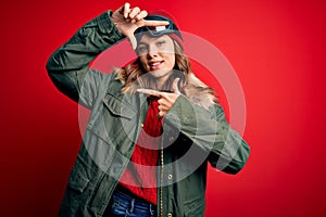 Young blonde girl wearing ski glasses and winter coat for ski weather over red background smiling making frame with hands and