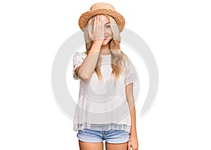 Young blonde girl wearing summer hat covering one eye with hand, confident smile on face and surprise emotion