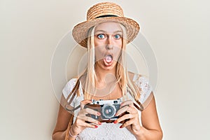 Young blonde girl wearing summer hat using camera afraid and shocked with surprise and amazed expression, fear and excited face