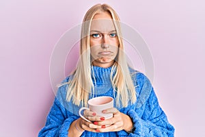 Young blonde girl wearing winter sweater and drinking a cup of hot coffee depressed and worry for distress, crying angry and