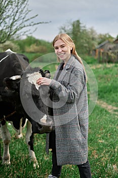 young blonde happy girl in coat hugs the cow in the green field