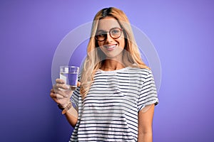 Young blonde healthy woman wearing glasses drinking glass of water over purple background with a happy face standing and smiling