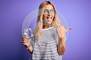 Young blonde healthy woman wearing glasses drinking glass of water over purple background pointing and showing with thumb up to