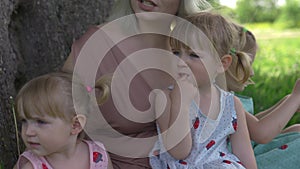 Young blonde hippie mother having quality time with her baby girls sitting and laughing under a tree in a park -