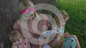 Young blonde hippie mother having quality time with her baby girls sitting and laughing under a tree in a park -