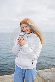 Young blonde lady stands near seaside and drink coffee, it is cold winter ourside, low temperature. White jacket and jeans