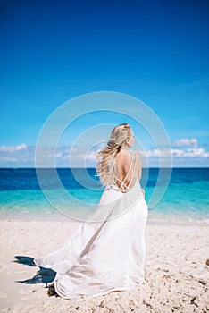 Young blonde long hair bride in long white dress with open back standing on the white sand beach.