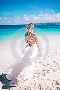 Young blonde long hair bride in white dress with open back standing on the beach. Tropical turquois ocean on the background.