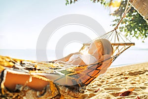 Young blonde longhaired woman relaxing in hammock hinged between palm trees on the sand beach