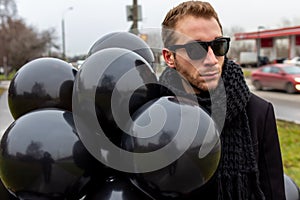 A young blonde man wearing sunglasses and black scarf holding black balloons, side view.