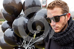 A young blonde man wearing sunglasses and black scarf holding black balloons, side view.