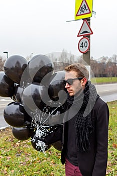 A young blonde man wearing sunglasses and black scarf holding black balloons, side view.