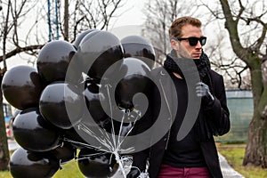 A young blonde man wearing sunglasses and black scarf holding black balloons, side view.