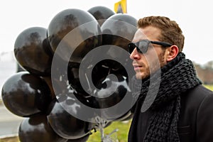 A young blonde man wearing sunglasses and black scarf holding black balloons, side view.