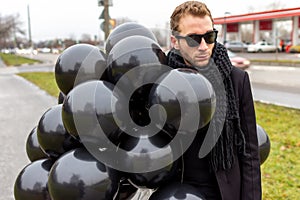 A young blonde man wearing sunglasses and black scarf holding black balloons, side view.