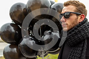 A young blonde man wearing sunglasses and black scarf holding black balloons, side view.