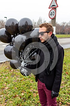 A young blonde man wearing sunglasses and black scarf holding black balloons, side view.