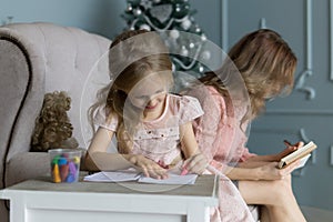 Young blonde mom sitting on couch in pink blouse writing plans. Christmas tree. Daughter raws pictures in the album with