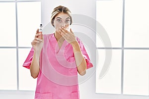 Young blonde nurse woman holding electronic cigarette covering mouth with hand, shocked and afraid for mistake