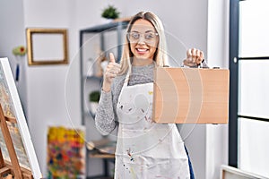 Young blonde painter woman holding wooden case at art studio smiling happy and positive, thumb up doing excellent and approval