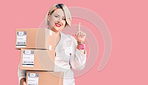 Young blonde plus size woman holding delivery cardboard package surprised with an idea or question pointing finger with happy