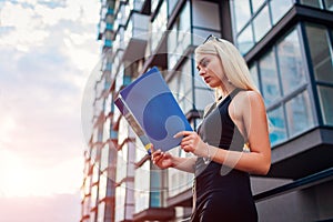 Young blonde real eastate agent reading contract by modern multi-storey building in city. Businesswoman examines project