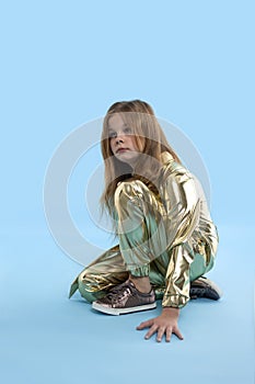 A  young blonde serious girl wearing in a golden jumpsuit sits on blue background looks aside and thinks about something. The