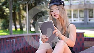 Young blonde sitting on bench working on tablet pc. Female college student using wireless internet in campus park