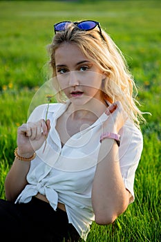 A young blonde is sitting in a clearing and holding a dandelion. Green grass.