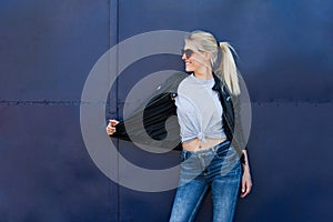 Young blonde smiling woman in casual grey t shirt sunglasses and black jacket against blue metal background outdoor in city t