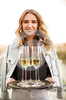 Young blonde smiling woman holding tray with champagne sparkling wine in flute glasses